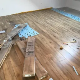 Układanie paneli podłogowych w jasnym pomieszczeniu na poddaszu. Widoczne paczki paneli 'Expert Choice Flooring', niebieska pianka izolacyjna, miarka i ołówek. Część paneli już ułożona, reszta...