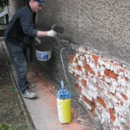 Mężczyzna w roboczych ubraniach nakłada zaprawę na odsłonięty fragment muru z czerwonej cegły, poniżej szorstkiej, jasnej elewacji budynku; obok stoi żółty opryskiwacz ciśnieniowy.