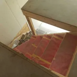 Wewnętrzna konstrukcja drewnianych schodów w trakcie budowy, widok z góry, zabezpieczone czerwoną folią, w tle fragment ściany i podłogi.