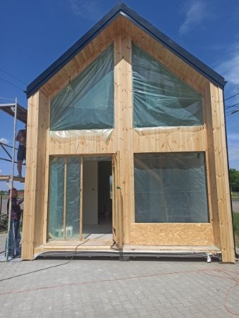 Surowy, drewniany dom w trakcie budowy z folią ochronną na oknach, widoczni pracownicy na rusztowaniu i podłączone kable elektryczne.