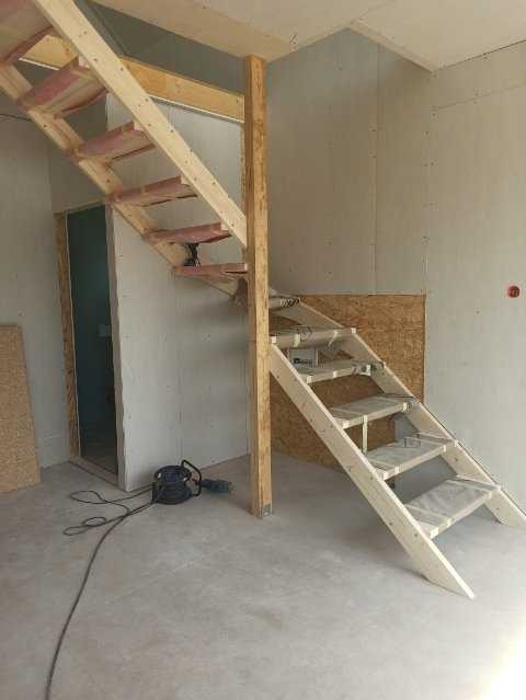 Wnętrze domu w trakcie budowy: drewniane schody modułowe bez balustrady, widoczne przewody elektryczne i narzędzia budowlane.