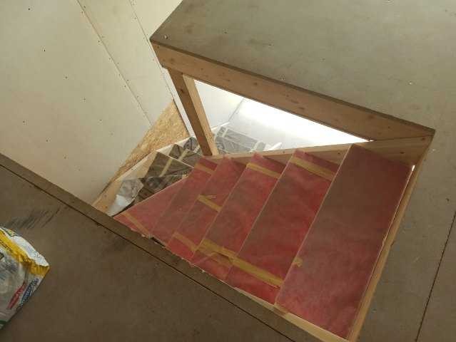 Wewnętrzna konstrukcja drewnianych schodów w trakcie budowy, widok z góry, zabezpieczone czerwoną folią, w tle fragment ściany i podłogi.