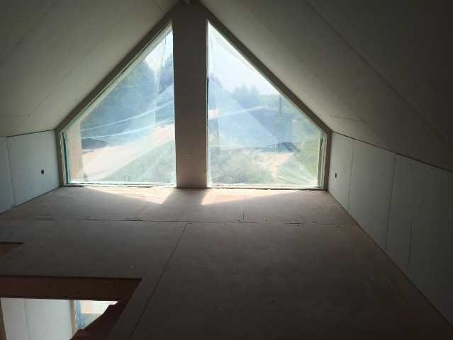 Wnętrze poddasza domu szkieletowego w trakcie budowy z dużym trójkątnym oknem i widoczną konstrukcją dachu.