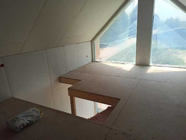 Wnętrze poddasza w trakcie budowy, widoczne okna trójkątne, drewniana konstrukcja podłogi i ściany wykończone płytami gipsowymi, worek materiałów budowlanych leżący na podłodze.