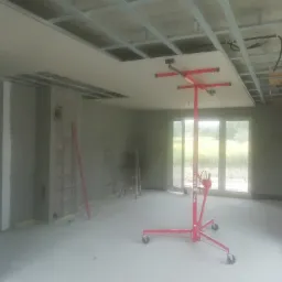 Wnętrze w trakcie montażu sufitu podwieszanego z płyt gipsowo-kartonowych, widoczny stelaż metalowy, płyta podtrzymywana przez podnośnik teleskopowy na kółkach, ściany w stanie surowym.
