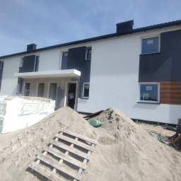 BUD-MAD ELEWACJE Sp. z o.o. - Dwukondygnacyjny budynek w trakcie prac elewacyjnych, widoczne ocieplenie styropianem oraz sterta piasku i paleta na pierwszym planie, fragment elewacji wykończony w kolorze grafitowym i imitacji...