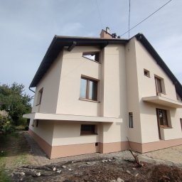 BUD-MAD ELEWACJE Sp. z o.o. - Dwukondygnacyjny dom z jasną elewacją i ciemnobrązowym dachem, widoczny z perspektywy żabiej, z fragmentem ogrodu i zaparkowanym busem w tle.