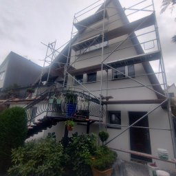 BUD-MAD ELEWACJE Sp. z o.o. - Dom jednorodzinny w trakcie prac elewacyjnych, widoczne rusztowania, świeżo pomalowana ściana oraz wiaderka z farbą przy ścianie.