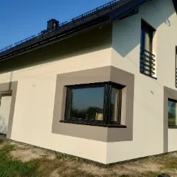 Nowoczesny dom jednorodzinny z ocieploną elewacją w odcieniach bieli i szarości, z czarnymi ramami okiennymi i dekoracyjnymi balustradami, uchwycony w słoneczny dzień.