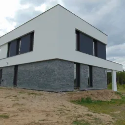 Nowoczesny dom z elewacją z szarego kamienia i białym tynkiem. Minimalistyczna bryła, duże okna z ciemnymi roletami. Surowe wykończenie elewacji.