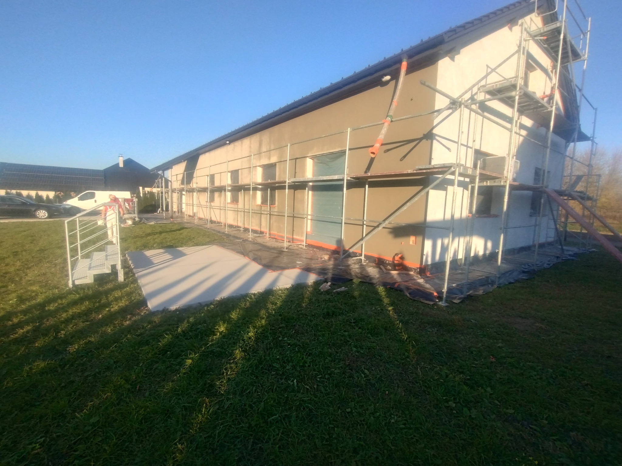 Dom jednorodzinny w trakcie prac elewacyjnych, z nałożoną warstwą tynku, otoczony rusztowaniem, z widocznymi elementami zabezpieczającymi okna i teren wokół budynku.