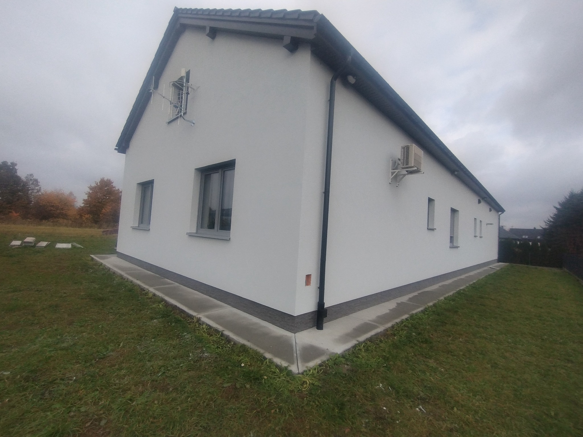 Boczny widok ocieplonego, białego domu z szarą stolarką okienną, anteną i klimatyzatorem na elewacji, z widocznym fragmentem trawnika i obrzeżem wokół budynku.