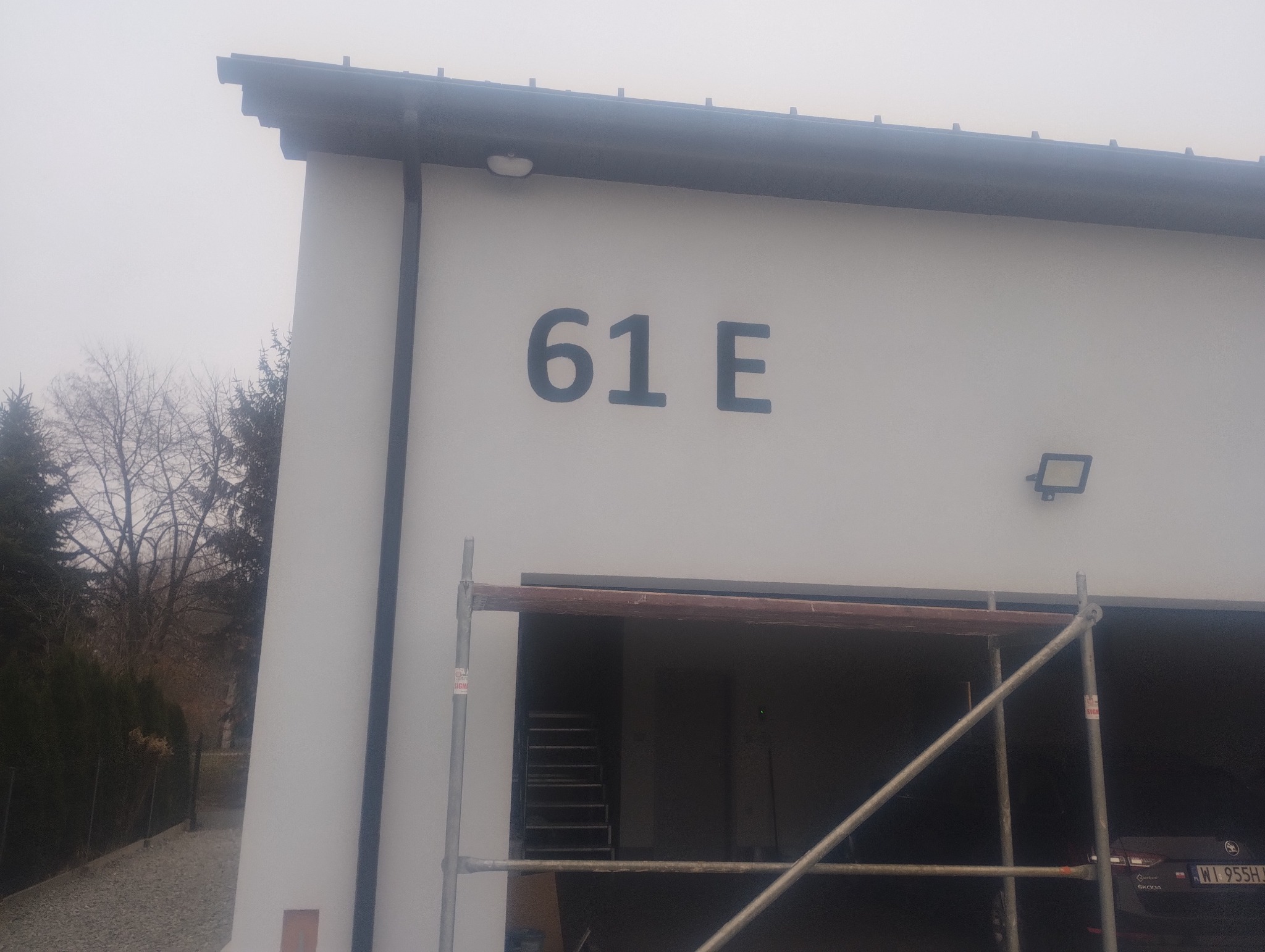 Biała elewacja budynku z numerem 61E, widoczne rusztowanie przed garażem, drzewa w tle, pochmurna pogoda.