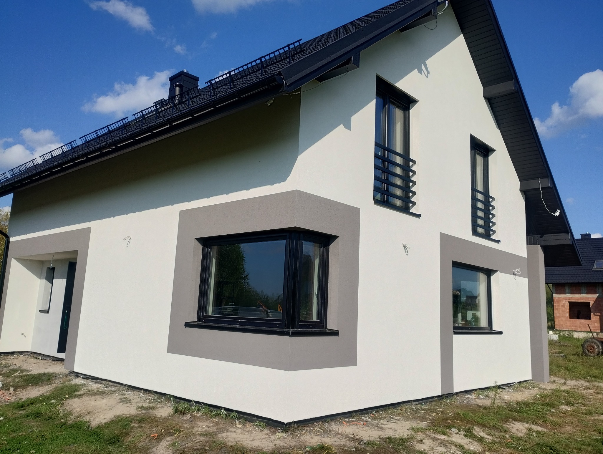 Nowoczesny dom jednorodzinny z białą elewacją i szarymi akcentami, czarnymi oknami i dachem, w słoneczny dzień.