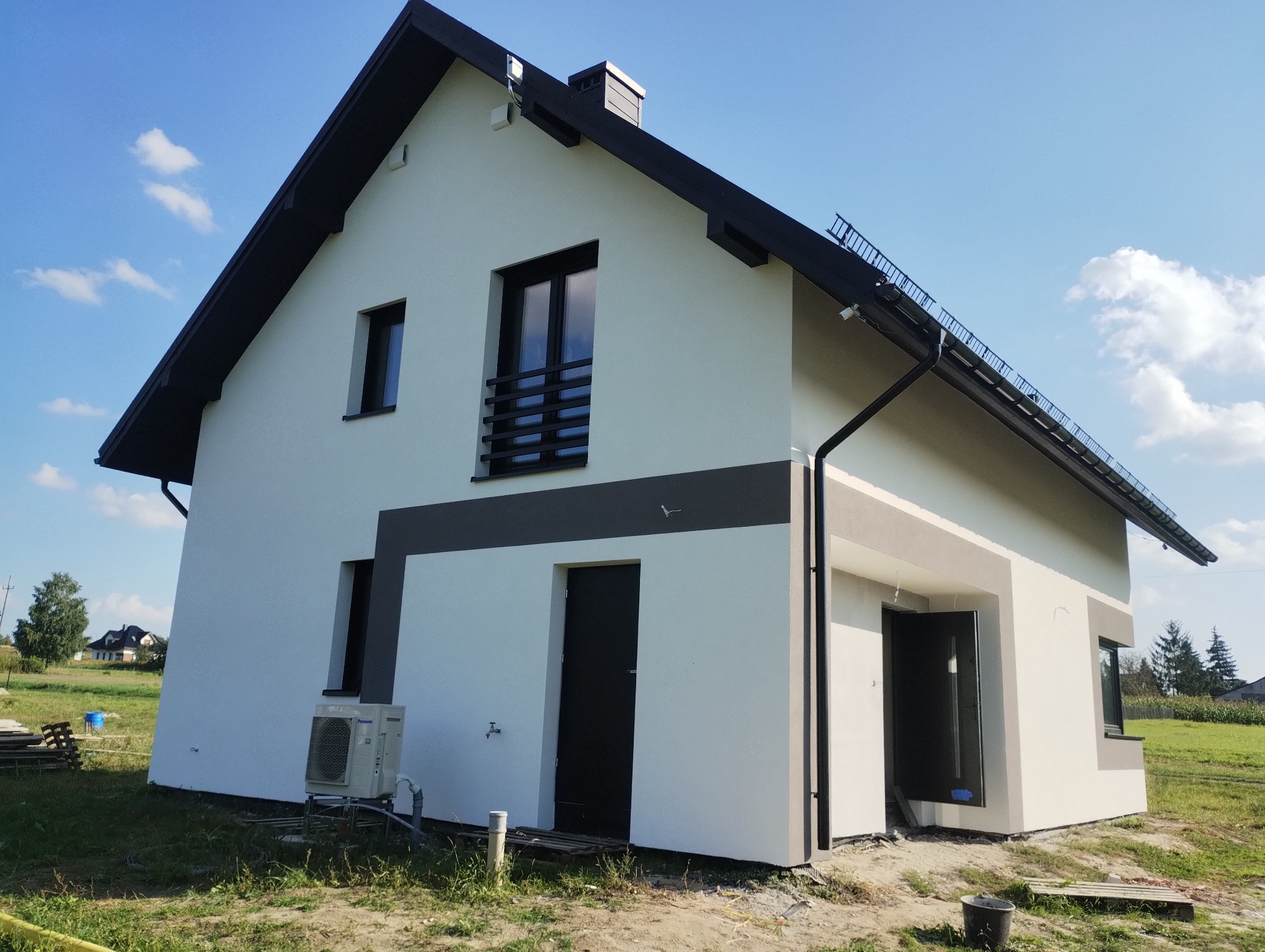 Nowoczesny dom jednorodzinny z białą elewacją i grafitowymi akcentami, widoczny klimatyzator przy ścianie, błękitne niebo w tle.