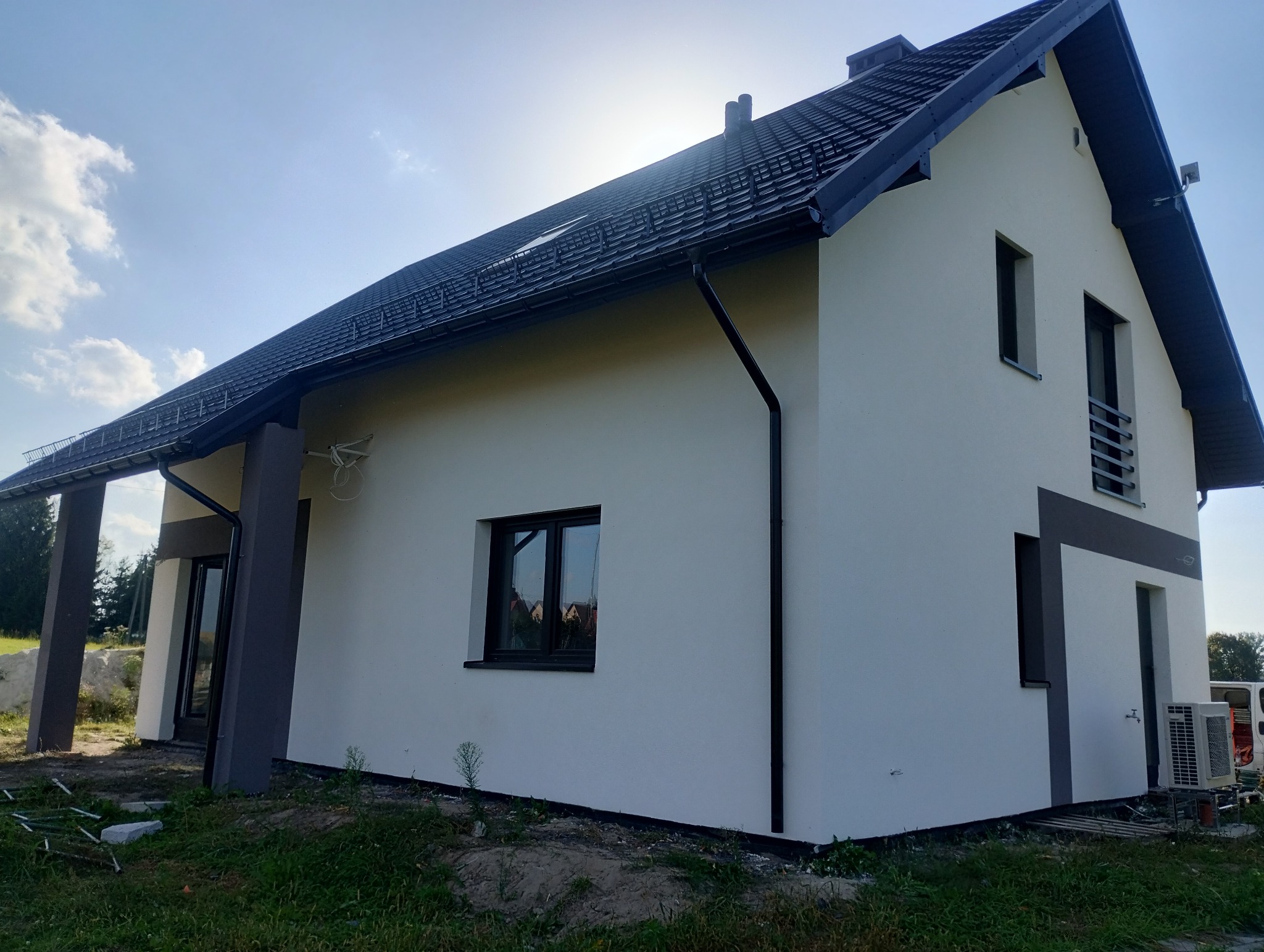 Dwukondygnacyjny dom jednorodzinny z jasną elewacją i ciemną dachówką, widoczny z boku pod kątem, z elementami dekoracyjnymi w kolorze grafitowym wokół okien i na filarach.