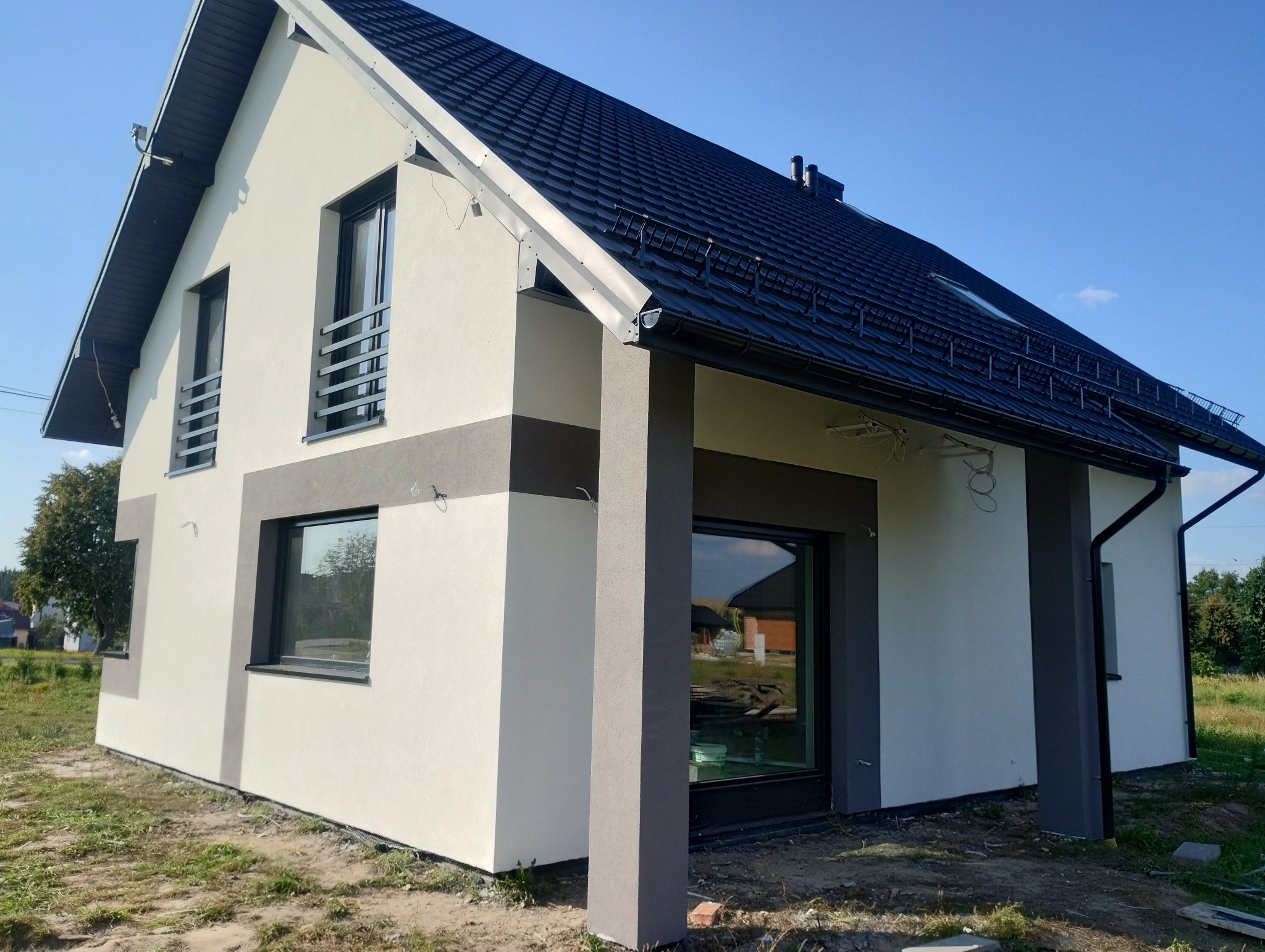 Nowoczesny dom jednorodzinny po ociepleniu elewacji, z elementami dekoracyjnymi w kolorze szarym, czarnym dachem i widocznymi przewodami elektrycznymi na ścianie.