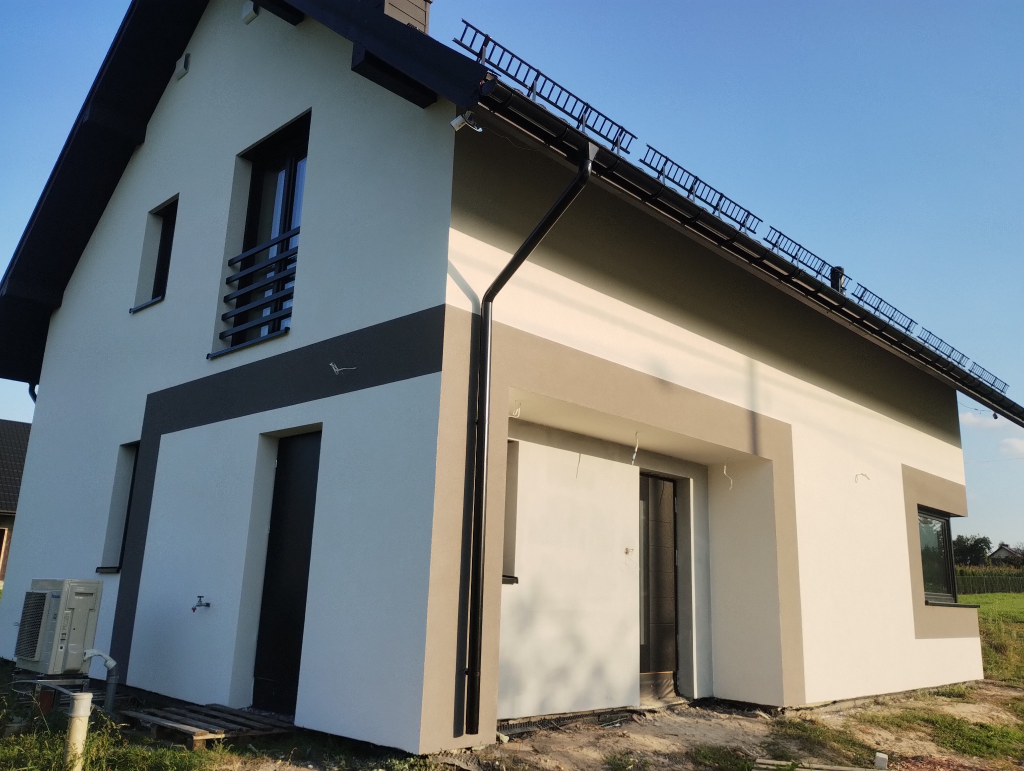 Nowoczesny dom jednorodzinny z jasną elewacją i grafitowymi elementami wykończeniowymi, widoczne orynnowanie i drabinka przeciwśniegowa na dachu, klimatyzator na zewnątrz budynku.