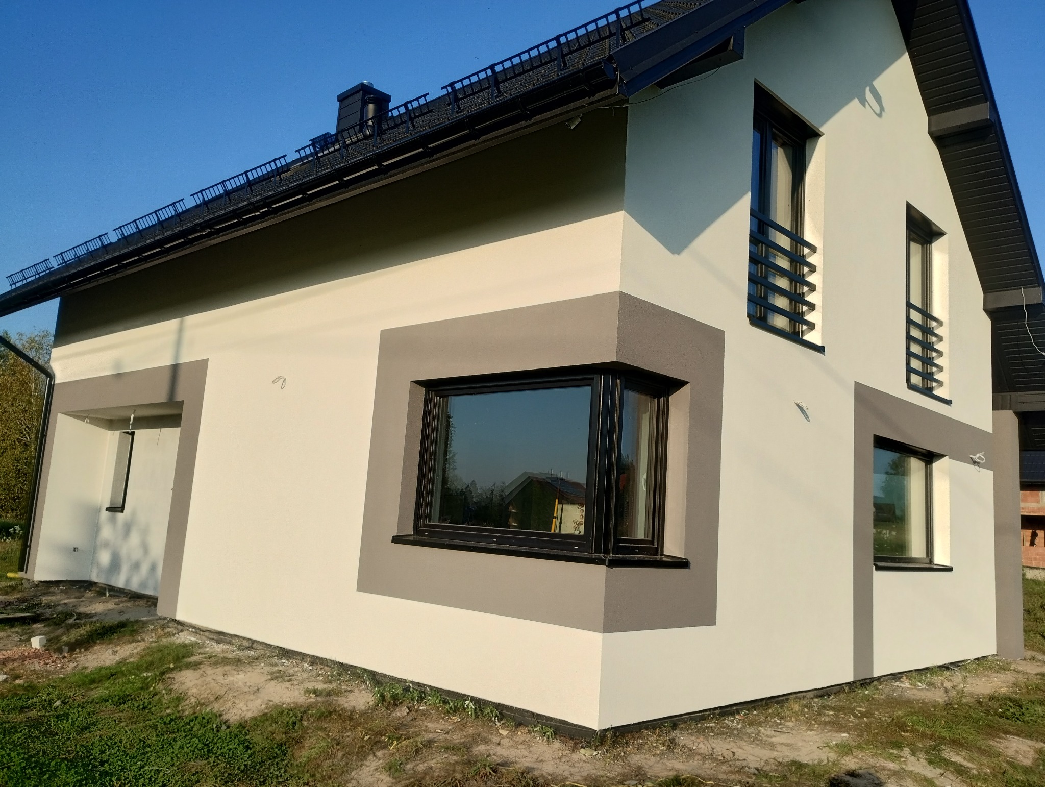 Nowoczesny dom jednorodzinny z ocieploną elewacją w odcieniach bieli i szarości, z czarnymi ramami okiennymi i dekoracyjnymi balustradami, uchwycony w słoneczny dzień.