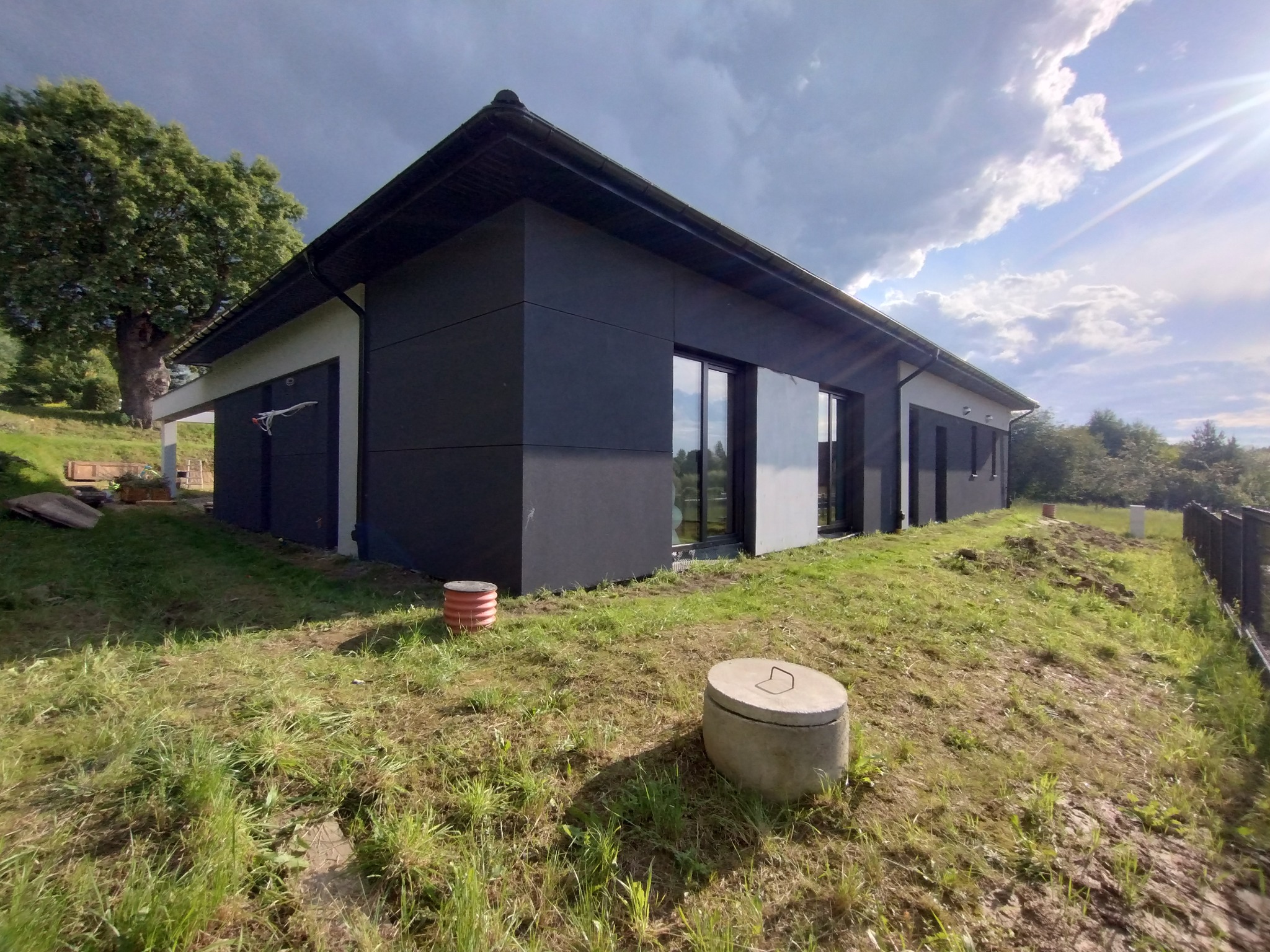 Nowoczesny dom jednorodzinny z ciemną elewacją panelową i dużymi oknami, widok z zewnątrz na nierównym terenie porośniętym trawą, z widocznymi elementami infrastruktury podziemnej.