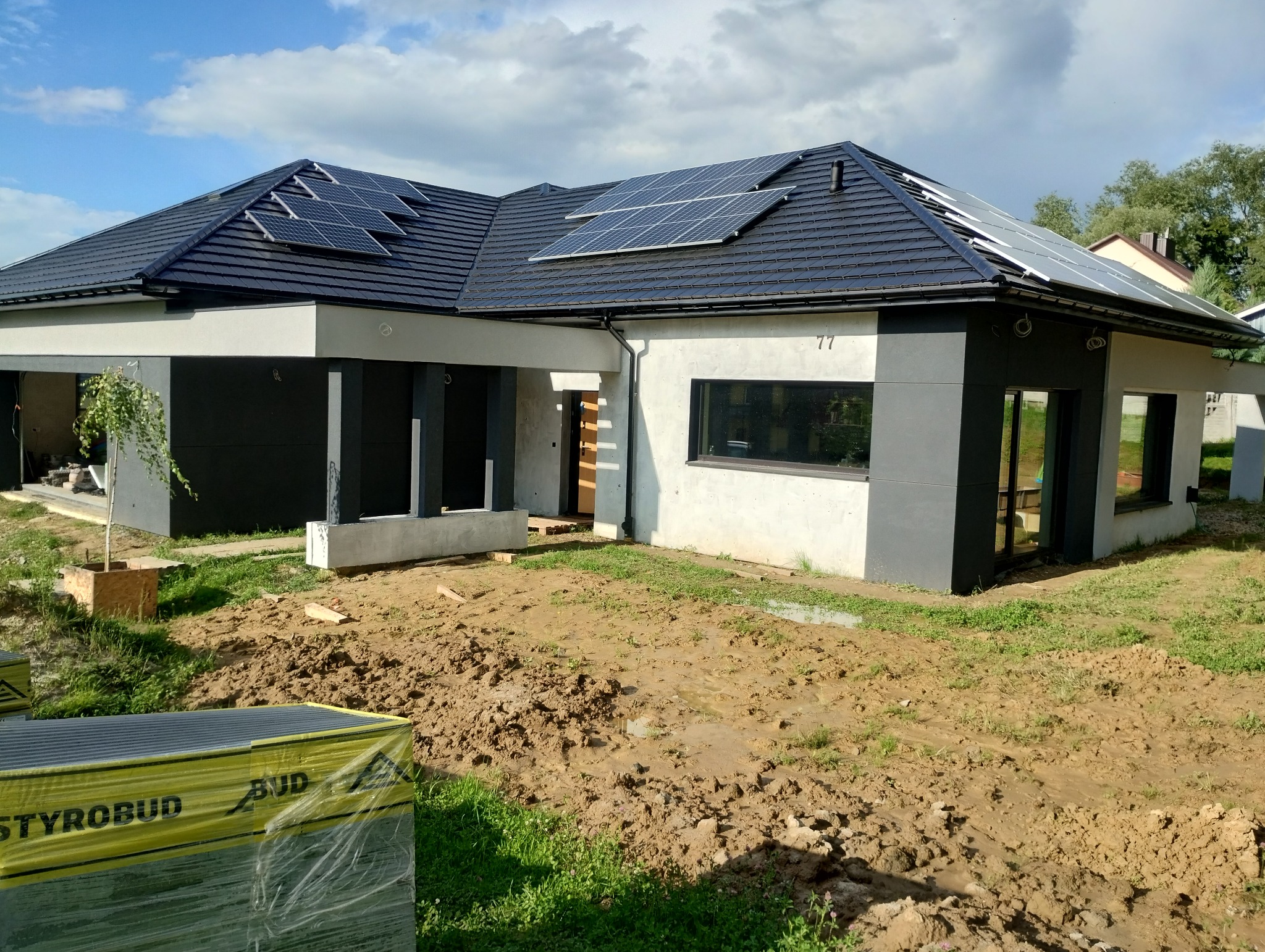 Nowoczesny dom jednorodzinny z panelami słonecznymi na dachu, w trakcie wykończenia elewacji, z widocznymi paczkami styropianu na pierwszym planie.