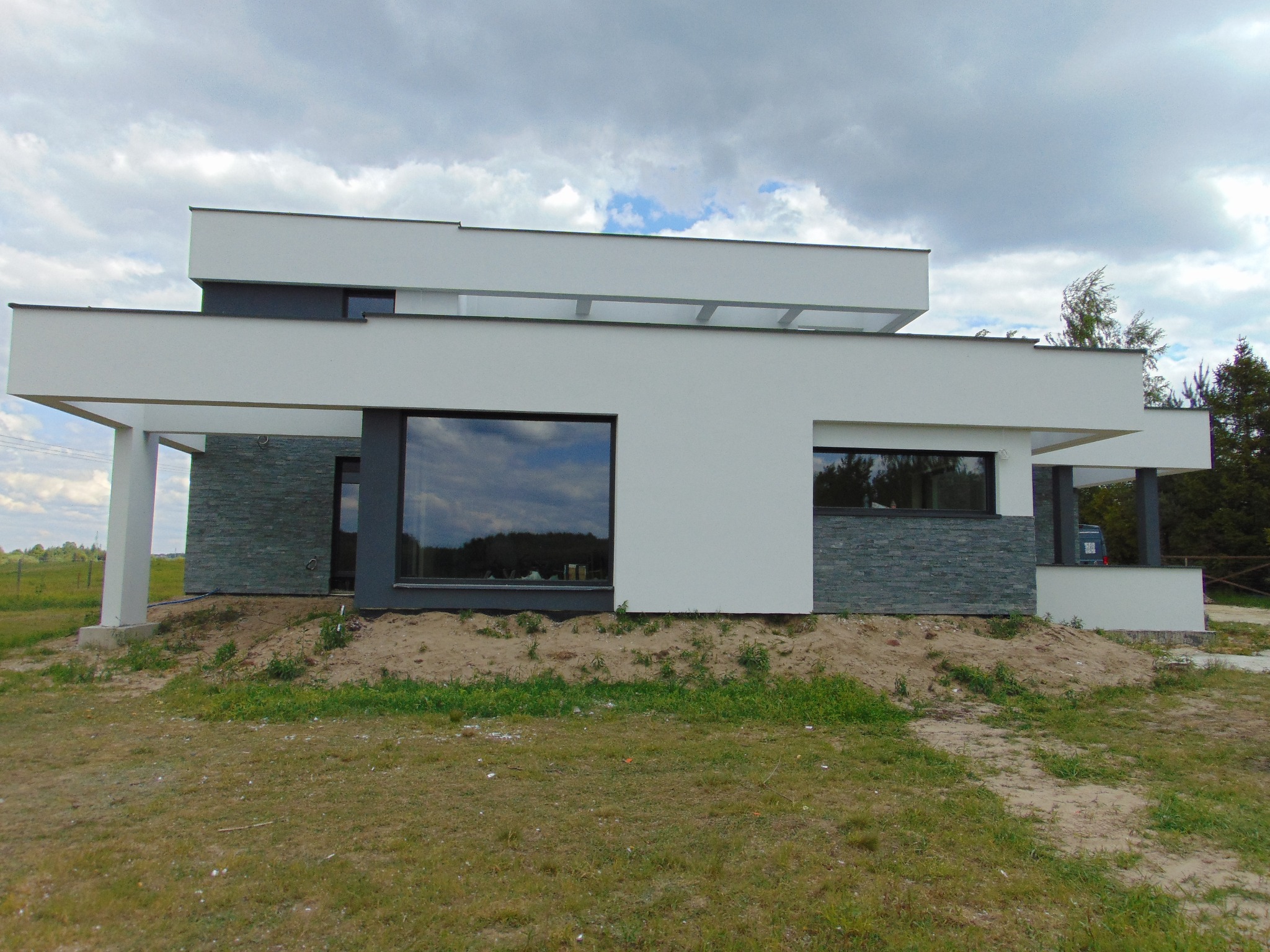 Nowoczesny dom z białą elewacją i szarym kamieniem dekoracyjnym. Duże okna odbijają krajobraz. Minimalistyczny design z płaskim dachem i geometrycznymi bryłami.