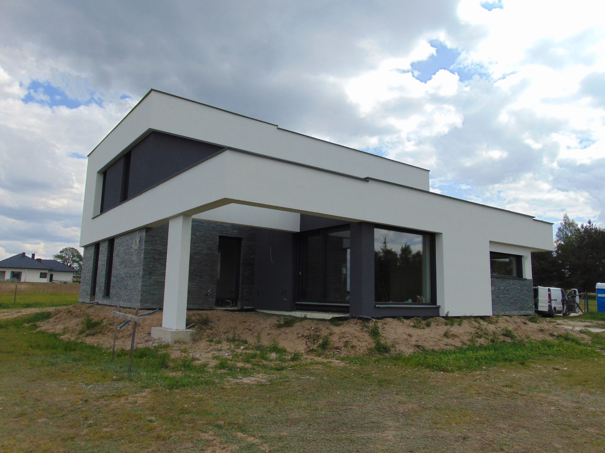 Nowoczesny dom z białą elewacją i szarym kamieniem dekoracyjnym, duże okna odbijające drzewa. Widoczne elementy wykończeniowe i otoczenie w trakcie aranżacji.