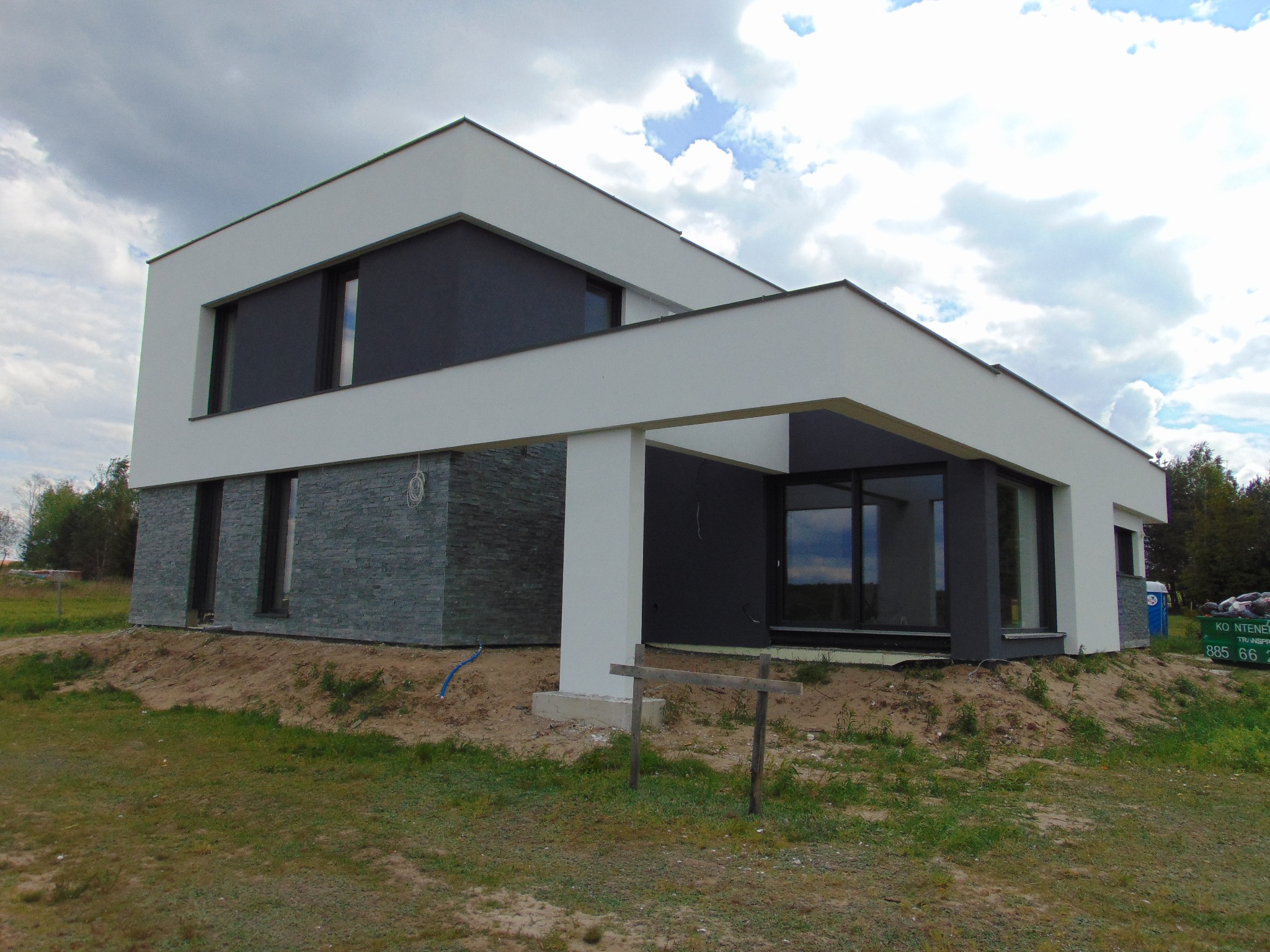 Nowoczesny dom z elewacją z szarego kamienia i białym wykończeniem, widok z zewnątrz w pochmurny dzień, z widocznym trawnikiem i elementami krajobrazu.