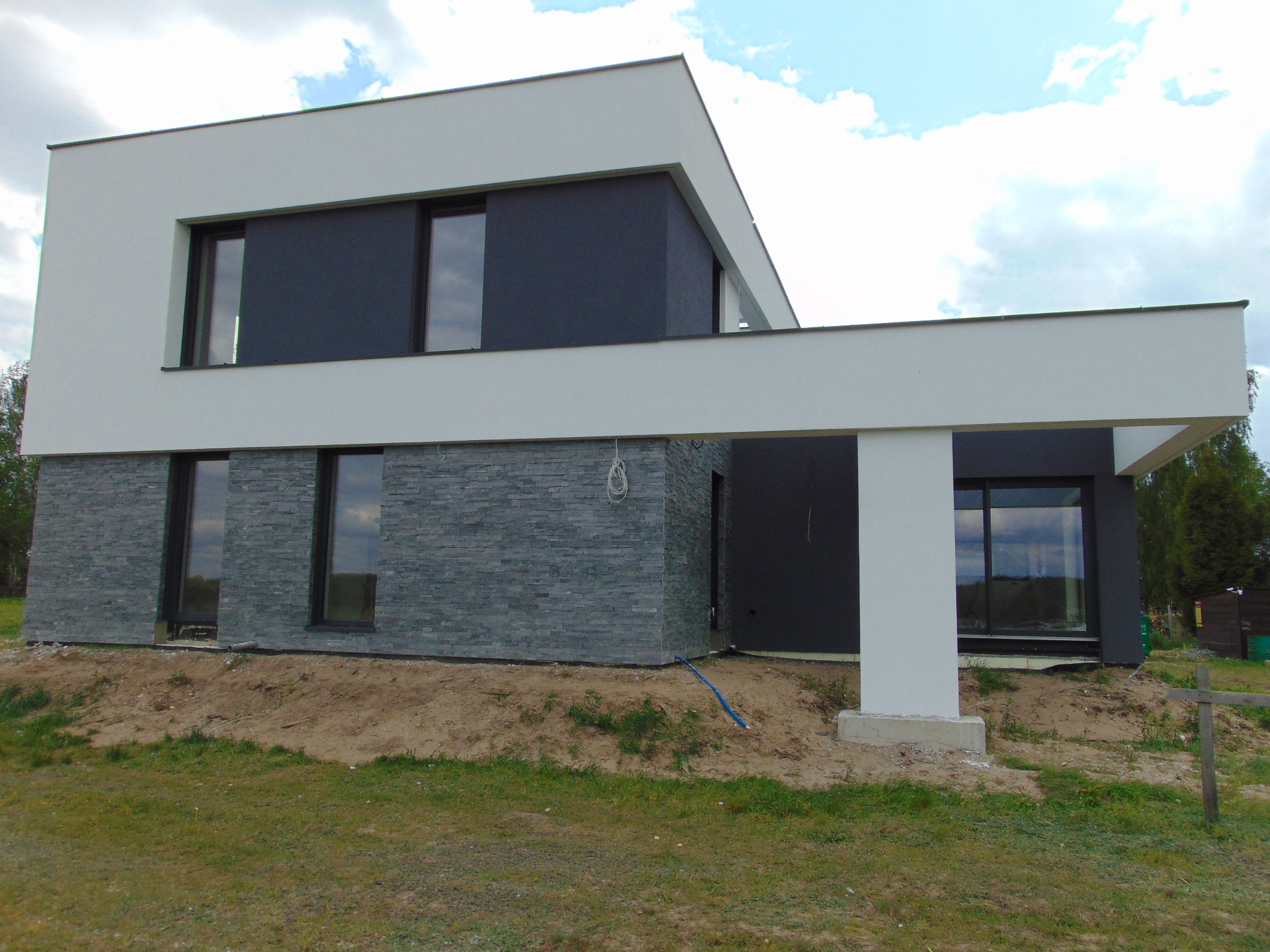 Nowoczesny dom z elewacją z szarej cegły i białym wykończeniem, okna z ciemnymi ramami, widok na trawnik i niebo z chmurami. Minimalistyczny design.