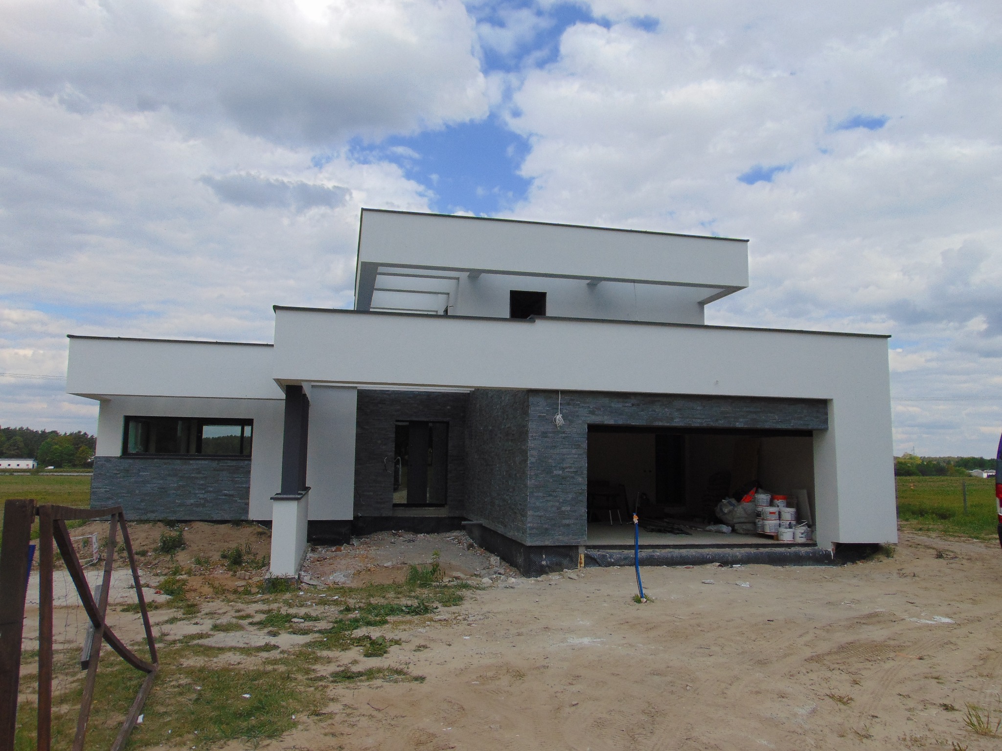 Nowoczesny dom jednorodzinny z elementami elewacji z kamienia dekoracyjnego, w trakcie wykańczania. Widoczne farby w garażu i teren wokół domu.