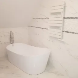 Nowoczesna, biała wanna wolnostojąca z chromowaną baterią w łazience wykończonej białym marmurem z dekoracyjnym pasem mozaiki i grzejnikiem drabinkowym.