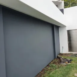 Świeżo pomalowana, gładka, szara elewacja budynku z nowoczesną, metalową bramą wejściową. W tle widoczny fragment trawnika z kamiennymi płytami i zwinięty wąż ogrodowy.