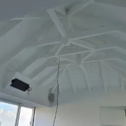 Białe drewniane belki stropowe w jasnym wnętrzu z widokiem na morze, w trakcie montażu oświetlenia i głośników.