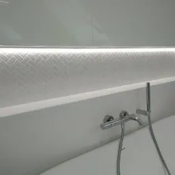 Biała łazienka z podświetlaną wnęką wyłożoną kafelkami w jodełkę, chromowana bateria wannowa z prysznicem ręcznym.