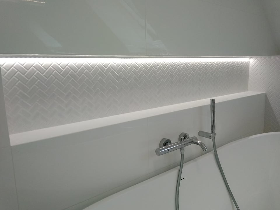 Biała łazienka z podświetlaną wnęką wyłożoną kafelkami w jodełkę, chromowana bateria wannowa z prysznicem ręcznym.