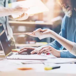 Zespół trzech osób pracuje przy biurku z laptopem i dokumentami, kobieta wskazuje ołówkiem na ekran, mężczyzna w tle trzyma dokumenty.