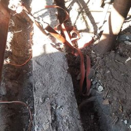 Adrian pele - Fragment rozkopanej ziemi z widocznym betonowym elementem zabezpieczonym pomarańczową taśmą transportową, metalowe podpory i kable w wykopie, cień drzewa na ziemi.