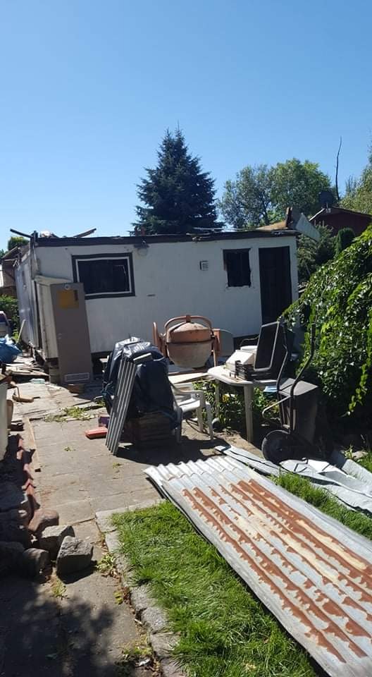 Uszkodzony budynek gospodarczy po rozbiórce, z widoczną betonową mieszarką, elementami konstrukcyjnymi i pofalowaną blachą na trawie.