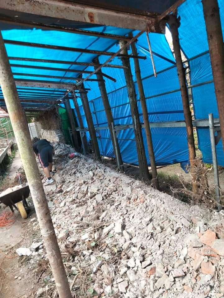 Demontaż konstrukcji osłoniętej niebieską plandeką, gruz i cegły na ziemi, widoczny pracownik i taczka.