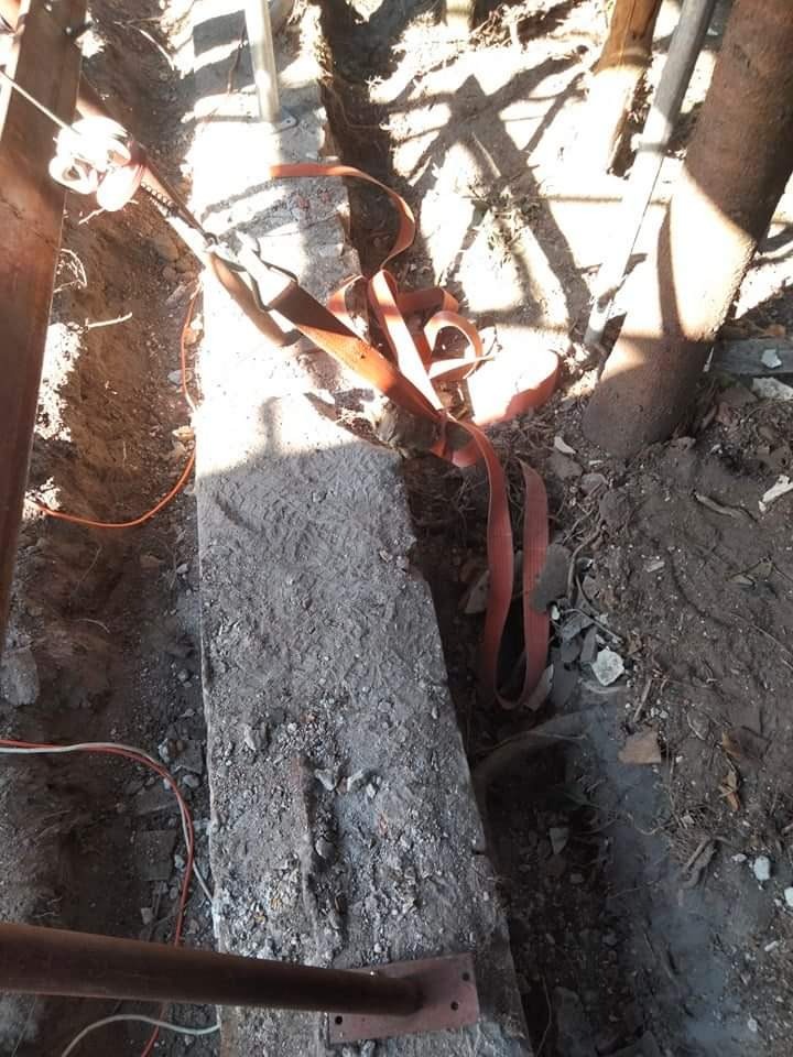 Fragment rozkopanej ziemi z widocznym betonowym elementem zabezpieczonym pomarańczową taśmą transportową, metalowe podpory i kable w wykopie, cień drzewa na ziemi.