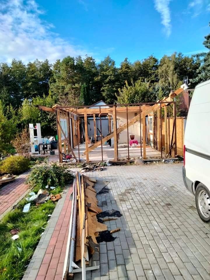 Częściowo rozebrany budynek z drewnianą konstrukcją, widoczne gruzy i elementy demontażu na brukowanym podjeździe, biały samochód dostawczy w tle, drzewa i błękitne niebo.