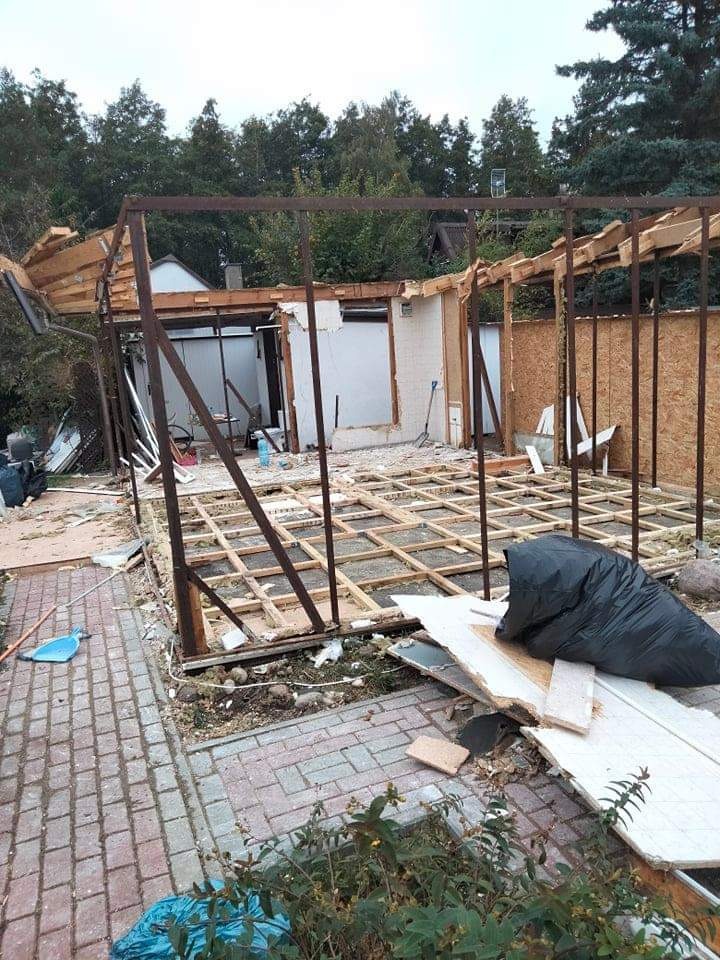 Częściowo rozebrany budynek z metalową konstrukcją szkieletową, widoczne elementy drewniane i płyty, otoczenie z kostki brukowej i roślinności, czarny worek na śmieci w pierwszym planie.