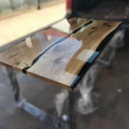 Stół z blatem z naturalnego drewna z żywicą epoksydową w kolorze niebieskim i czarnym, widoczne słoje drewna i nieregularne krawędzie, nogi stołu owinięte folią ochronną.