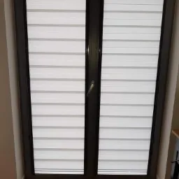 Zamknięte okno dwuskrzydłowe z szarymi ramami i zamontowanymi roletami w biało-szare poziome pasy, widoczne drewniane panele podłogowe.