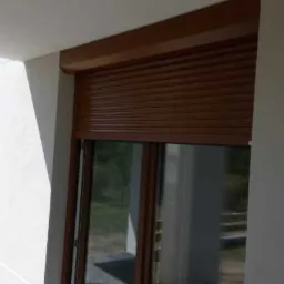 Brązowa roleta zewnętrzna częściowo opuszczona na oknie nowoczesnego domu z białymi ścianami.