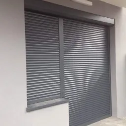 Zewnętrzna roleta antywłamaniowa w kolorze grafitowym, częściowo opuszczona, zamontowana na oknie i drzwiach w nowoczesnym budynku o jasnej elewacji, oświetlona g&oacute;rnym światłem.