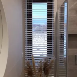 Białe poziome listwy w oknie łazienki, z widokiem na zimowy krajobraz, dekorowane suchymi trawami w wazonie.