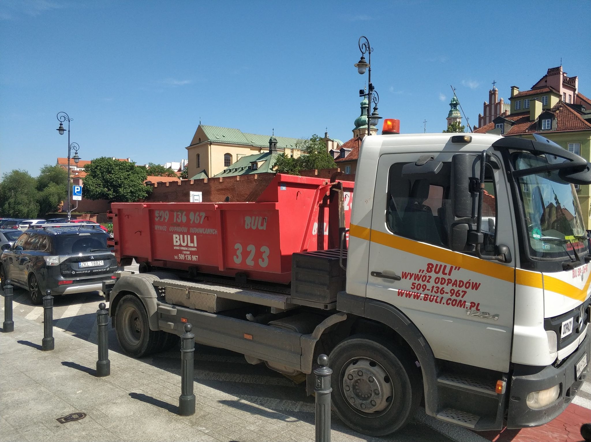 Ciężarówka z czerwonym kontenerem na platformie, zaparkowana przy chodniku z ozdobnymi słupkami, w tle zabytkowa zabudowa Warszawy.