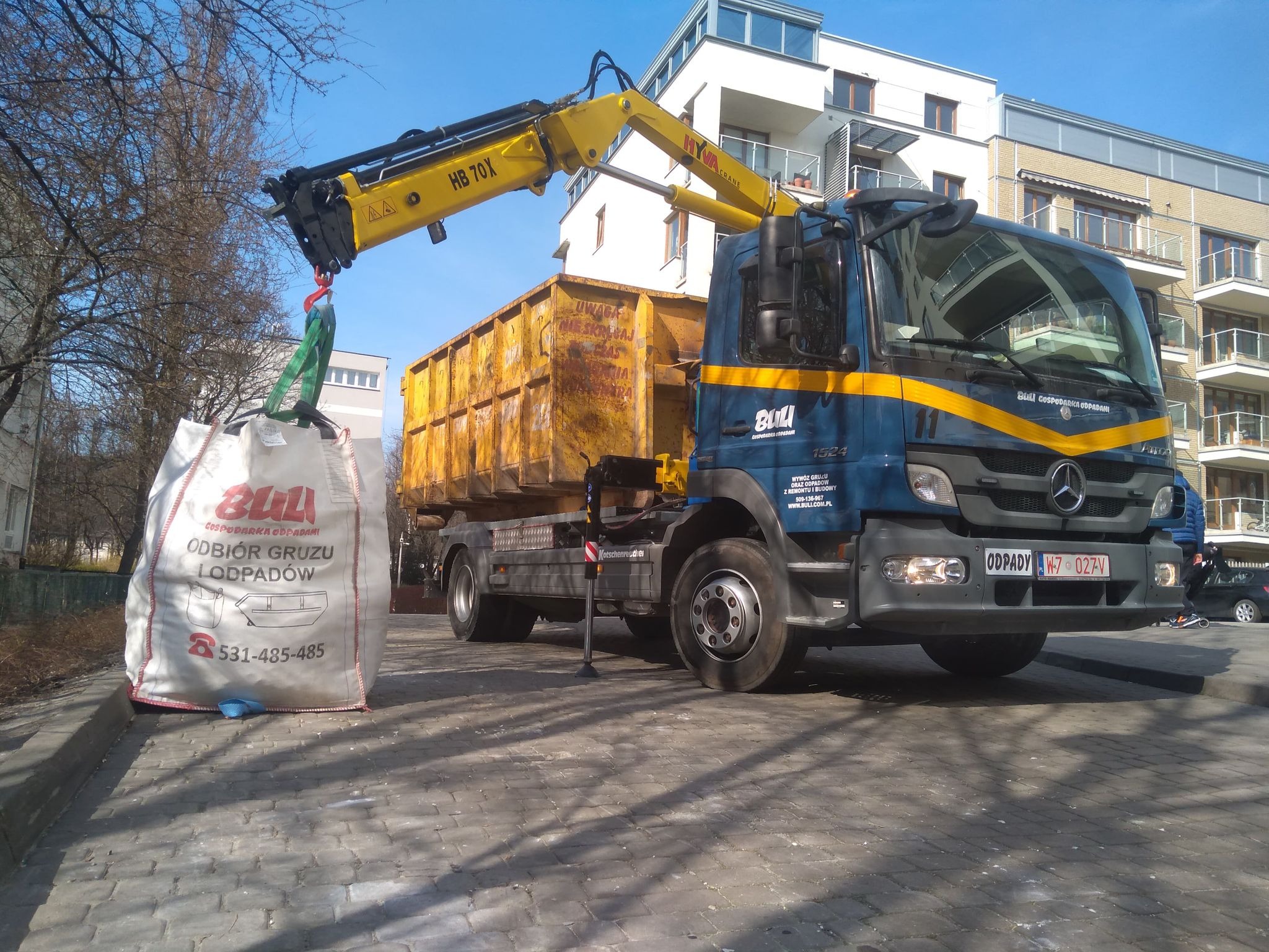 Ciężarówka z żółtym kontenerem i dźwigiem podnosi duży, biały worek z napisem 'Odbiór gruzu i odpadów', na tle nowoczesnego budynku mieszkalnego.