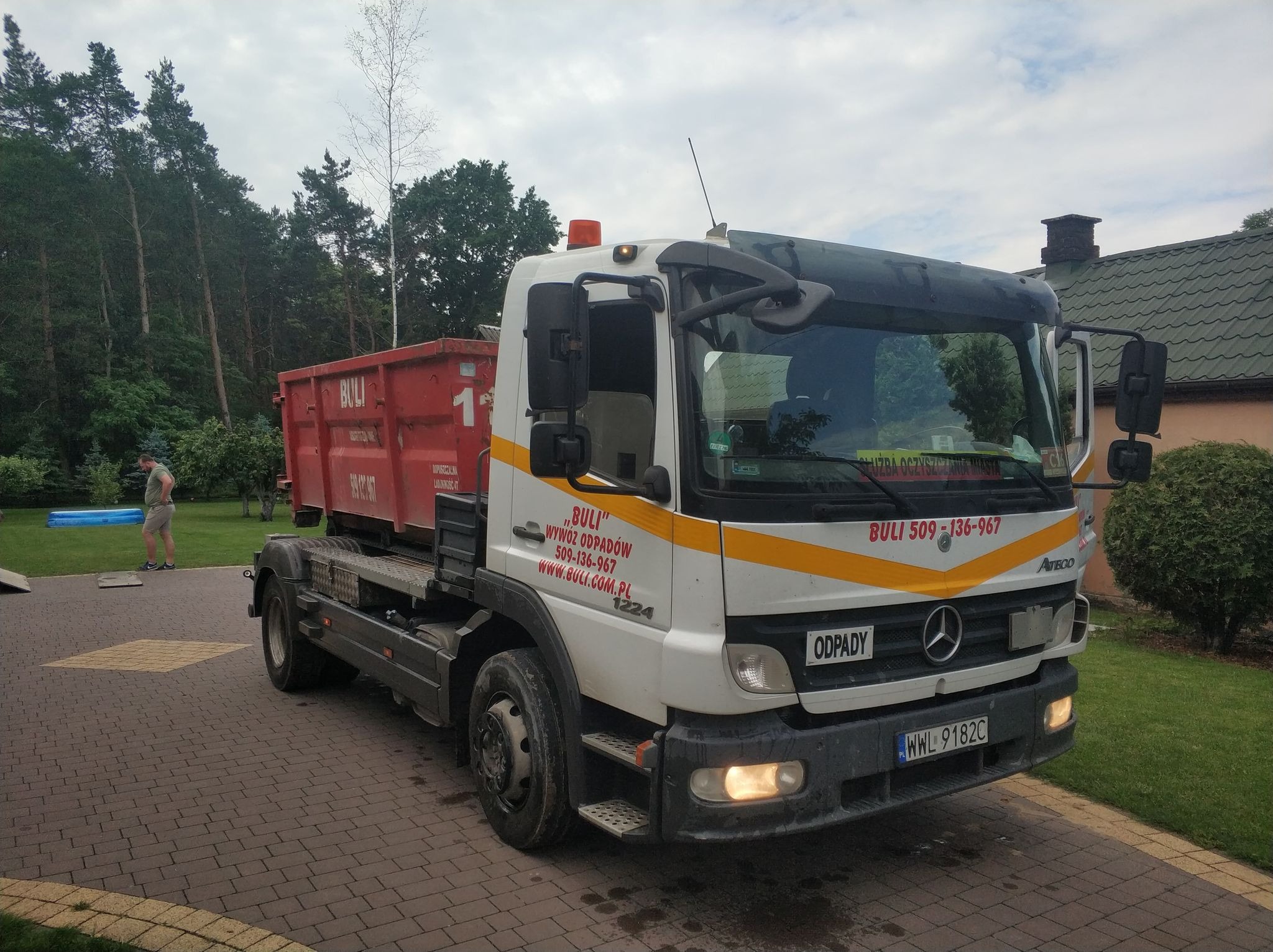 Ciężarówka Mercedes Atego z czerwonym kontenerem na platformie, zaparkowana na kostce brukowej przed domem z zielonym dachem, w tle drzewa i osoba przy basenie.